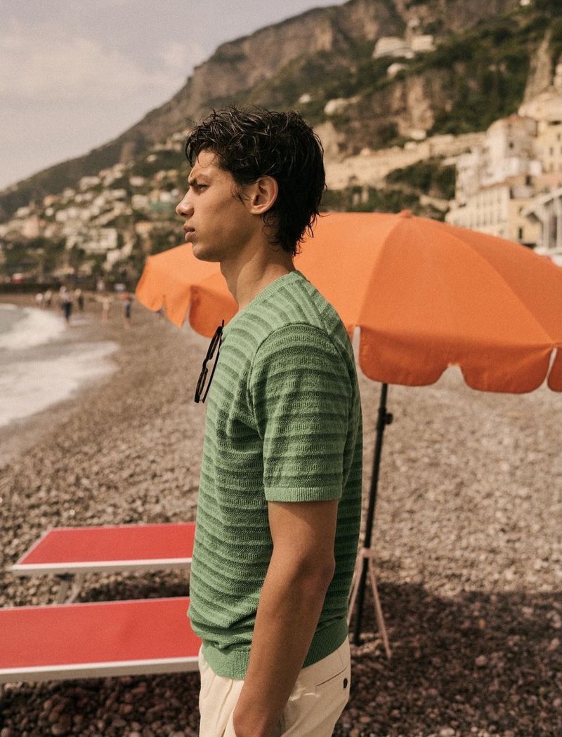 Young man stands on pebble beach wearing green striped shirt, with mountain town in background on sunny day by orange umbrella.