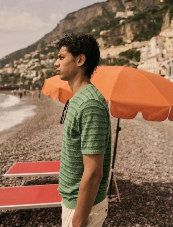 Young man stands on pebble beach wearing green striped shirt, with mountain town in background on sunny day by orange umbrella.