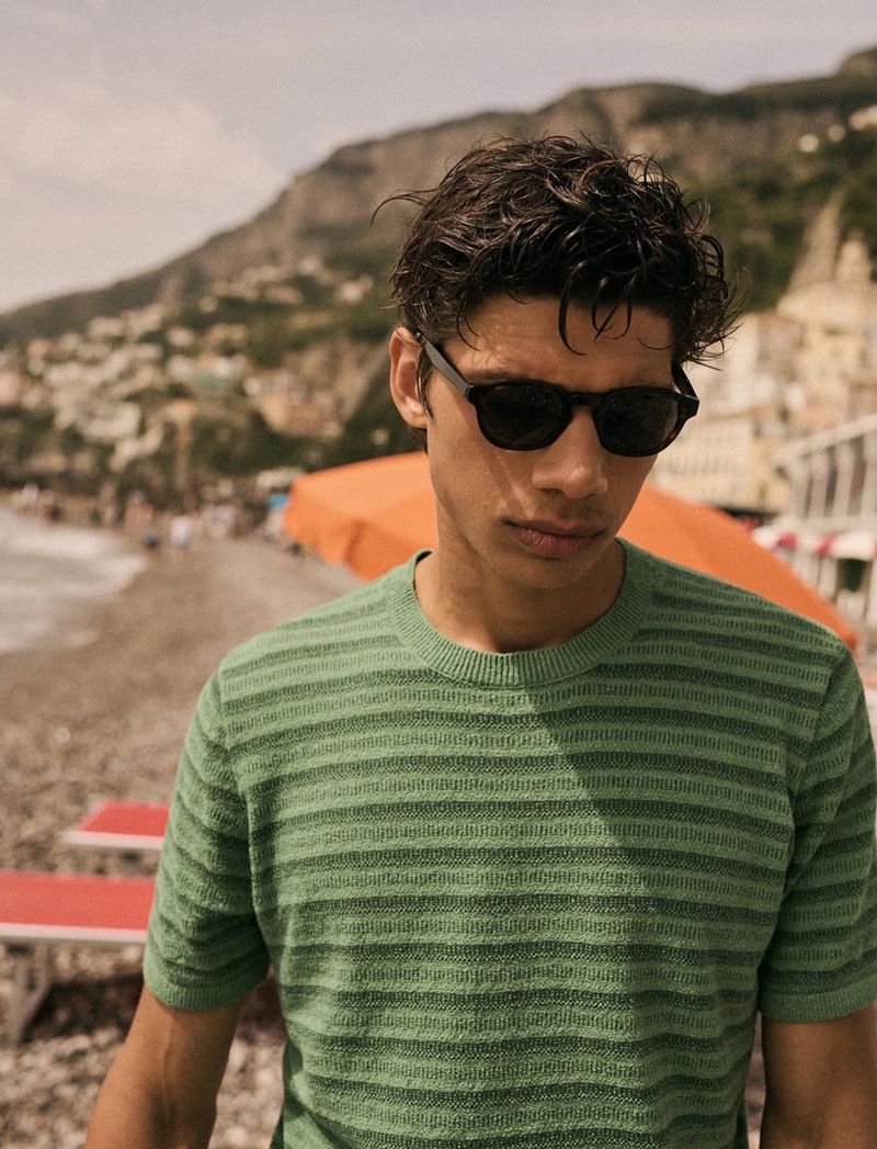 Close-up of a young adult male in a striped green shirt and sunglasses standing on a beach with a mountain backdrop.