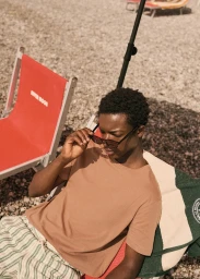 A Black man wearing sunglasses and a camel t-shirt sitting on a beach chair with a red canvas seat on a pebbled beach.