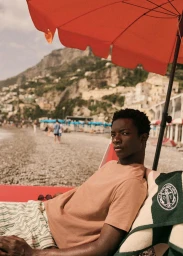A young Black man seated under a red beach umbrella wearing a peach t-shirt and striped shorts on a rocky coast.