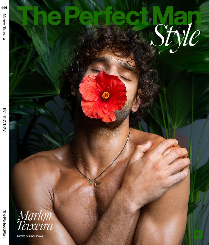 Shirtless man with curly hair poses with a red hibiscus flower covering his mouth, against a backdrop of vibrant green tropical foliage.