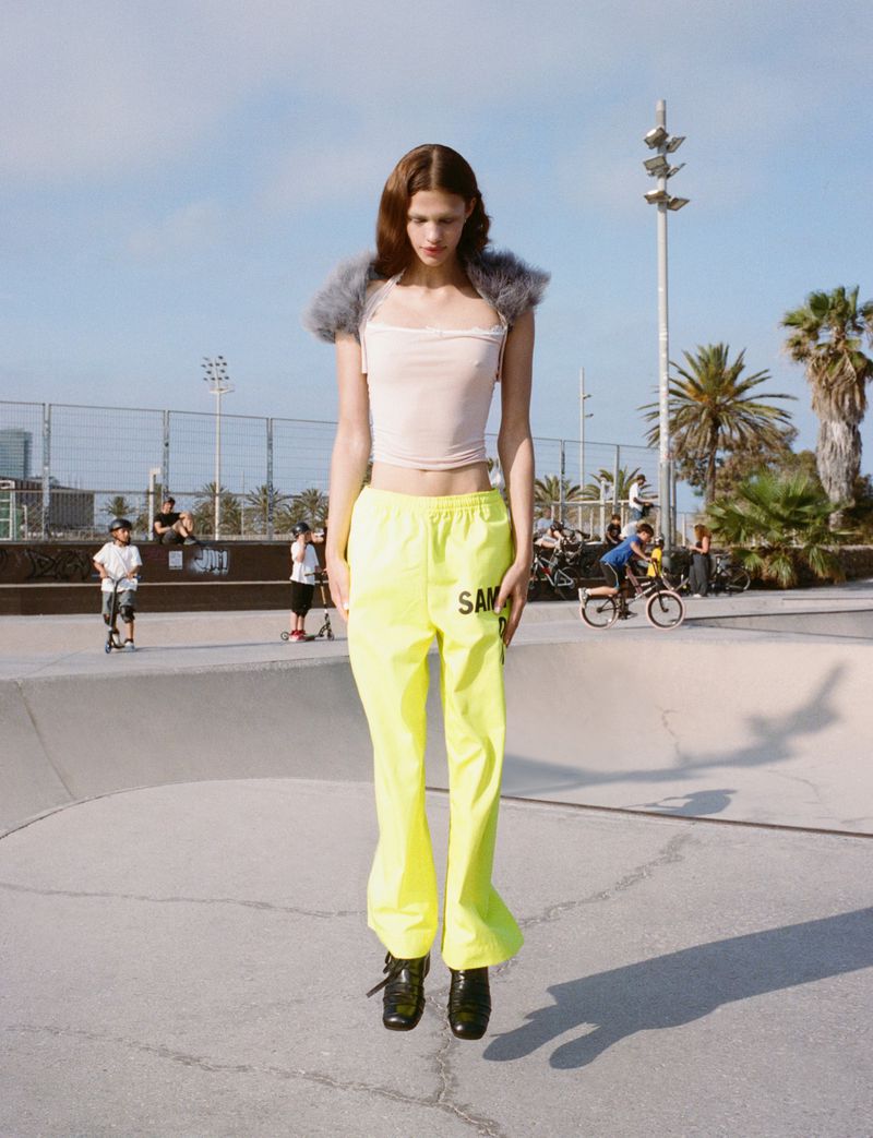 A young woman stands in a skate park wearing neon yellow pants, a pale pink top, and black shoes on a sunny day with a blue sky.