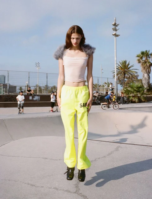 A young woman stands in a skate park wearing neon yellow pants, a pale pink top, and black shoes on a sunny day with a blue sky.