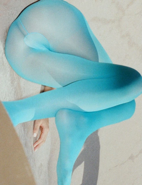 Woman's legs and torso twisted and bent in light blue sheer hosiery against a neutral surface and lit by soft, diffused light.