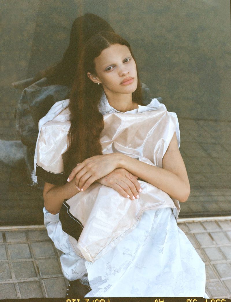 Fashion shot of a young woman with long brown hair wearing a plastic top and white skirt, seated on a tiled floor in natural light.