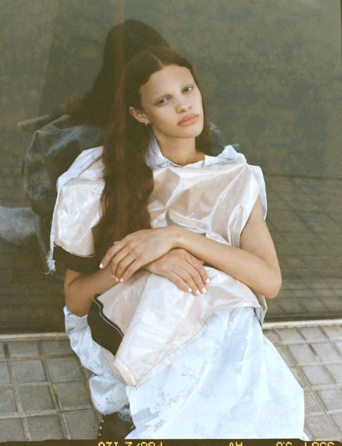 Fashion shot of a young woman with long brown hair wearing a plastic top and white skirt, seated on a tiled floor in natural light.