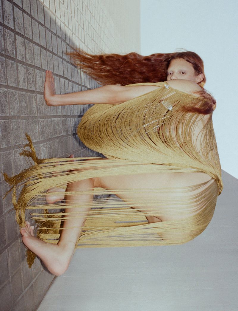 A redhead woman is wrapped in a gold fringed shawl against a grey tiled wall and a light grey wall with an unusual perspective.
