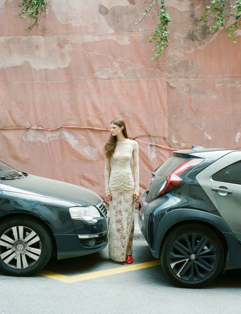 Adult female fashion model poses between two cars in long beige lace dress and red shoes against a pink wall with green vines.