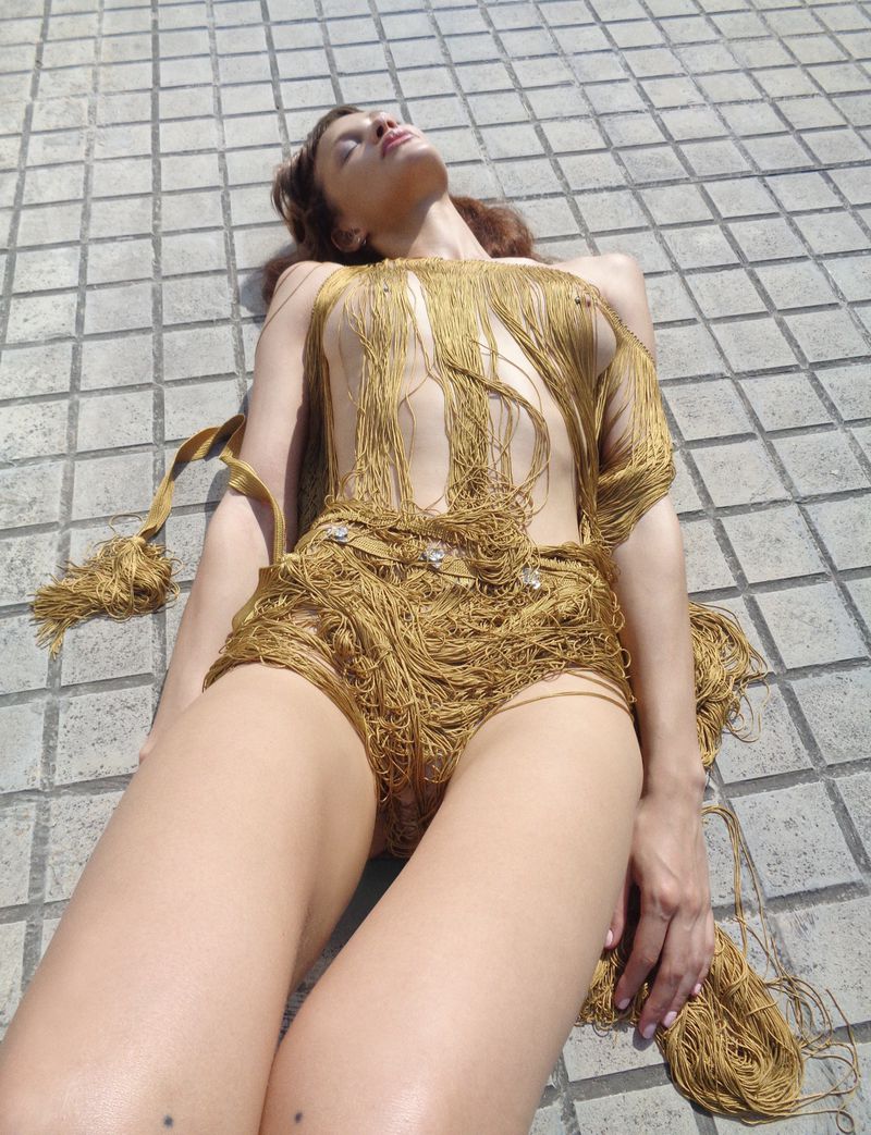 Woman lying on paved surface wearing fringe outfit in sunlight, eyes closed, fashion-forward pose. Brown hair, light-skinned, outdoor environment.