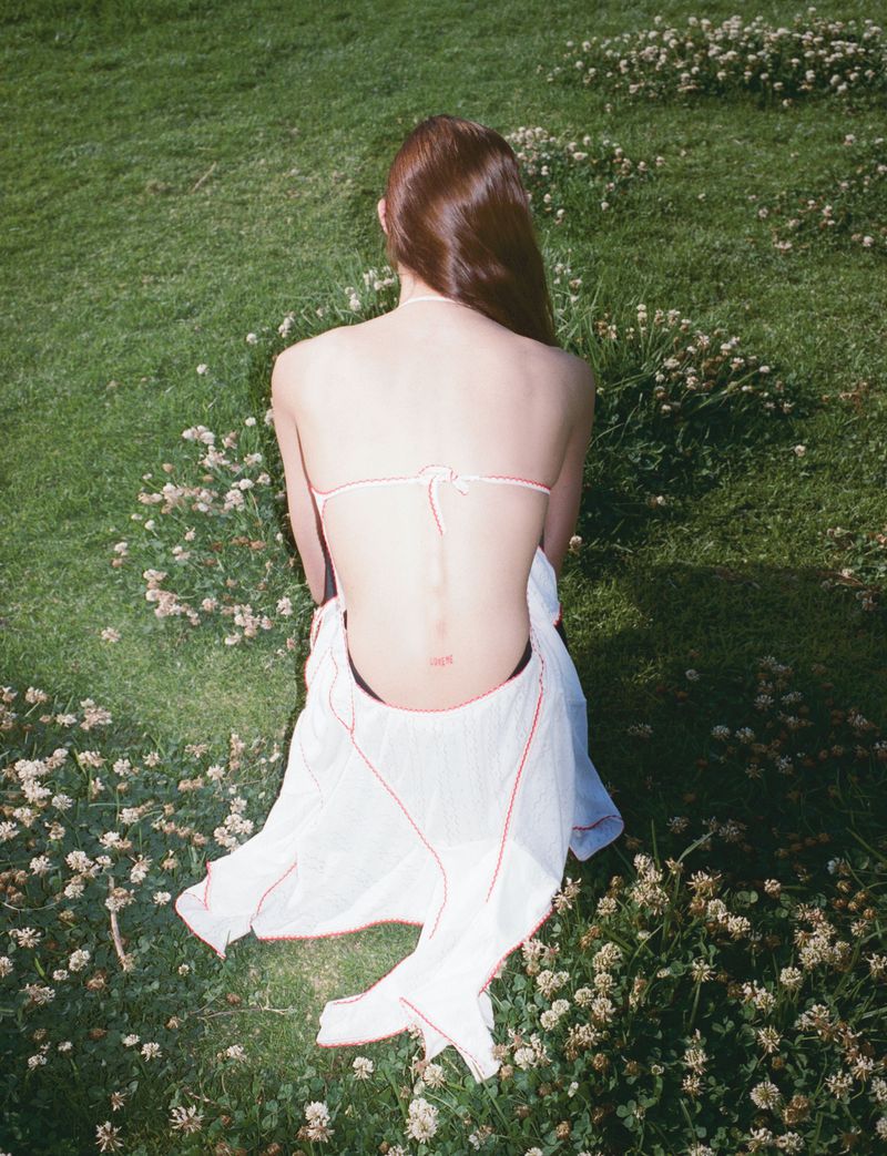 Back view of a woman in a bikini top and white cover-up sitting in a grassy field with clovers on a sunny day, natural lighting.