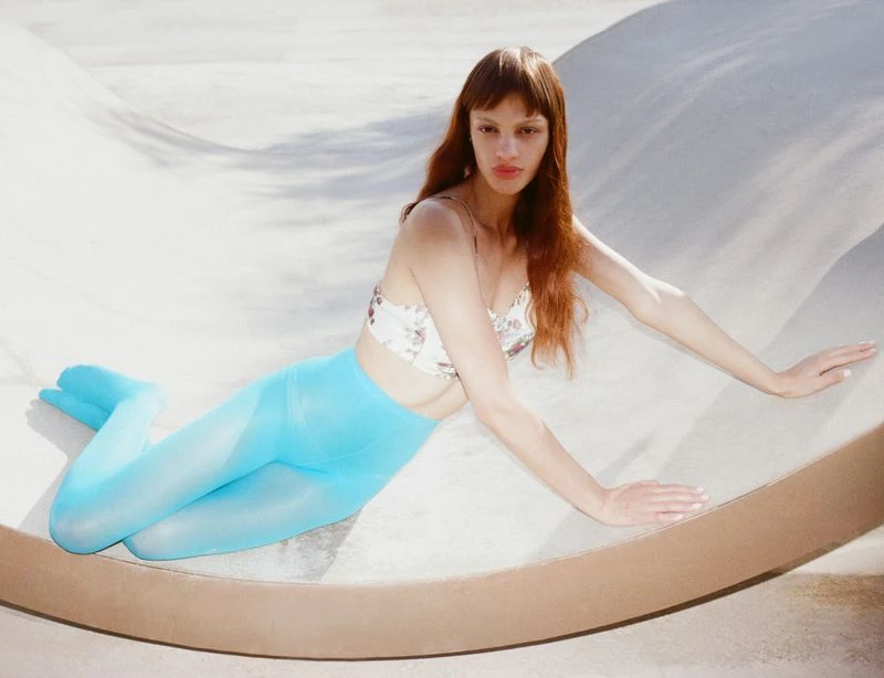 A woman with long auburn hair poses in a floral top and blue tights while reclining in a concrete skateboarding bowl, lit by bright sunlight.