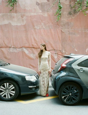 Model standing between two parked cars against a weathered pink wall for Urban Solstices in Hube Magazine