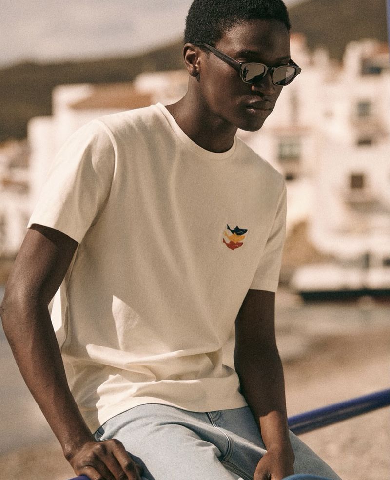 Portrait of an adult Black male model wearing a cream T-shirt and sunglasses sitting outdoors against a backdrop of coastal buildings.