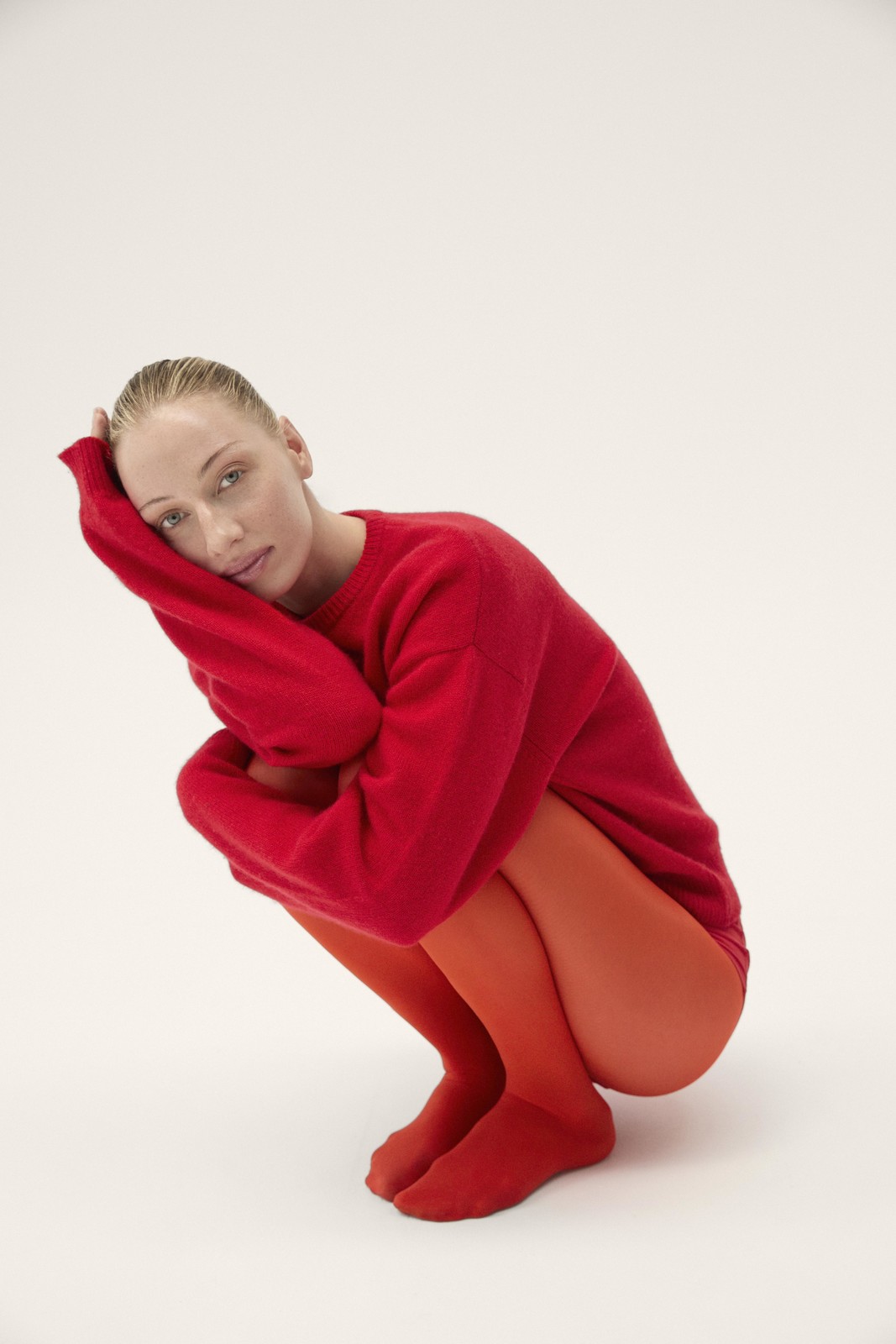 A blond-haired young adult female model in a red sweater and tights crouches in a studio with a neutral background, gazing at the camera.