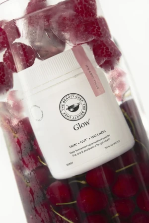 Product shot of The Beauty Chef Glow powder in a glass jar with ice cubes, raspberries, and cherries against a white backdrop.