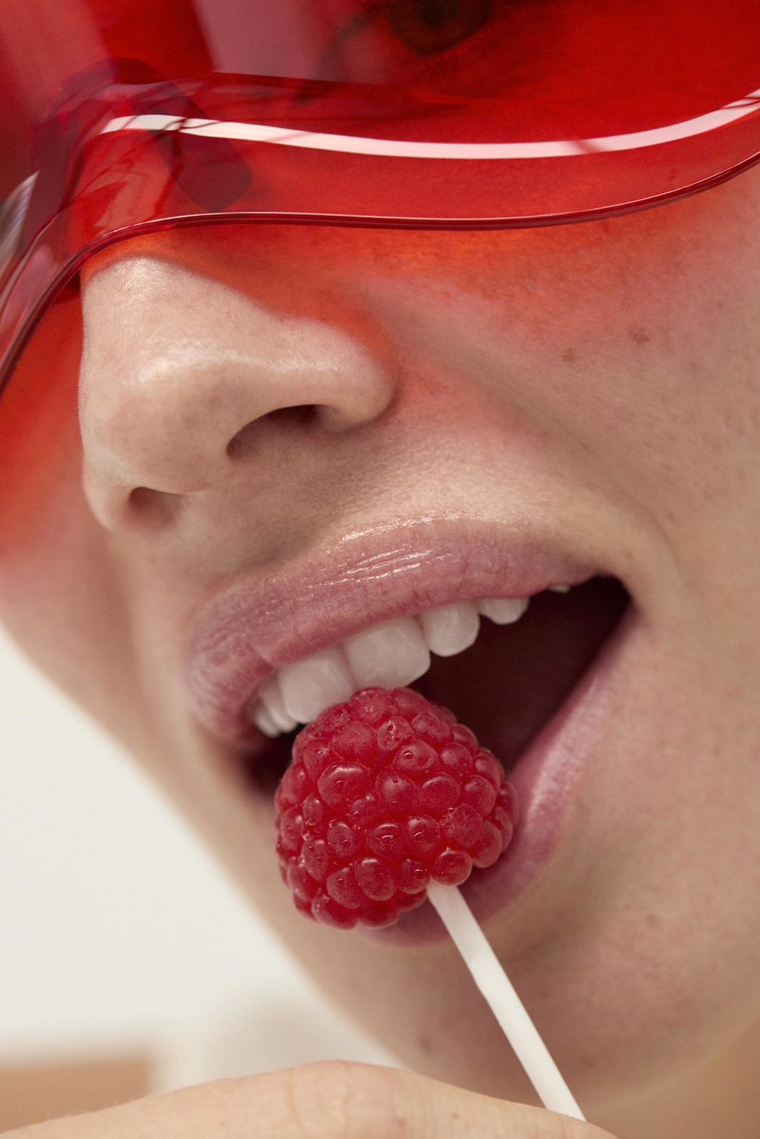 Close-up of a person with red glasses eating a raspberry lollipop, focusing on the mouth, teeth, and candy details against white background.