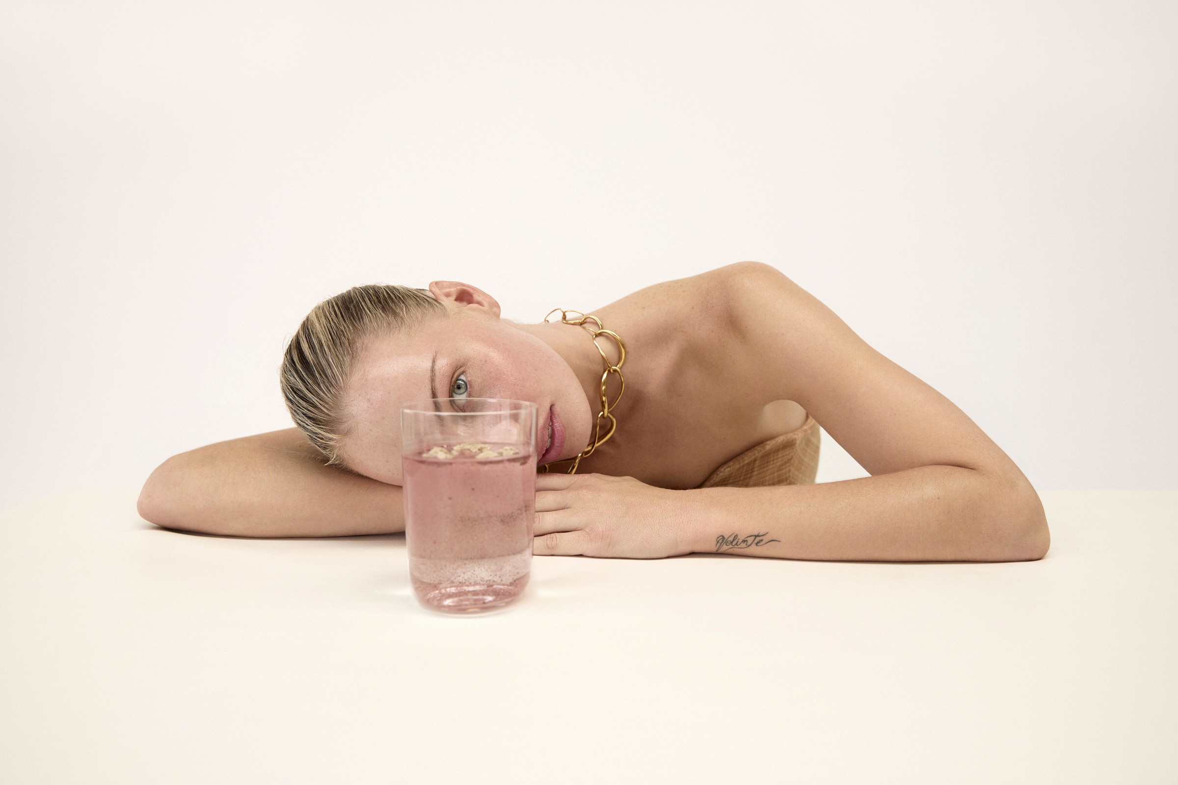 Attractive blonde woman lounges next to a glass of rosé-colored water against a plain backdrop, complemented by a stylish gold chain.