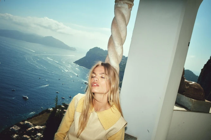 Woman posing in yellow top with a coastal view featuring mountains and turquoise sea, captured on a sunny day with architectural elements.