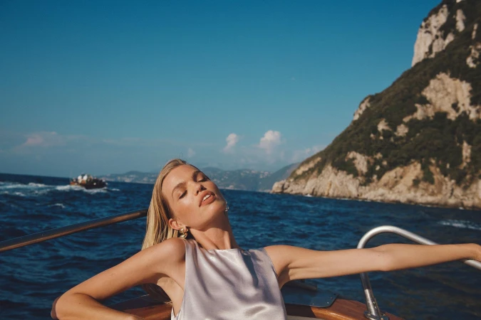 A blonde woman in a silver top relaxes on a boat with eyes closed against a backdrop of blue sea, sky, and a lush green mountain.