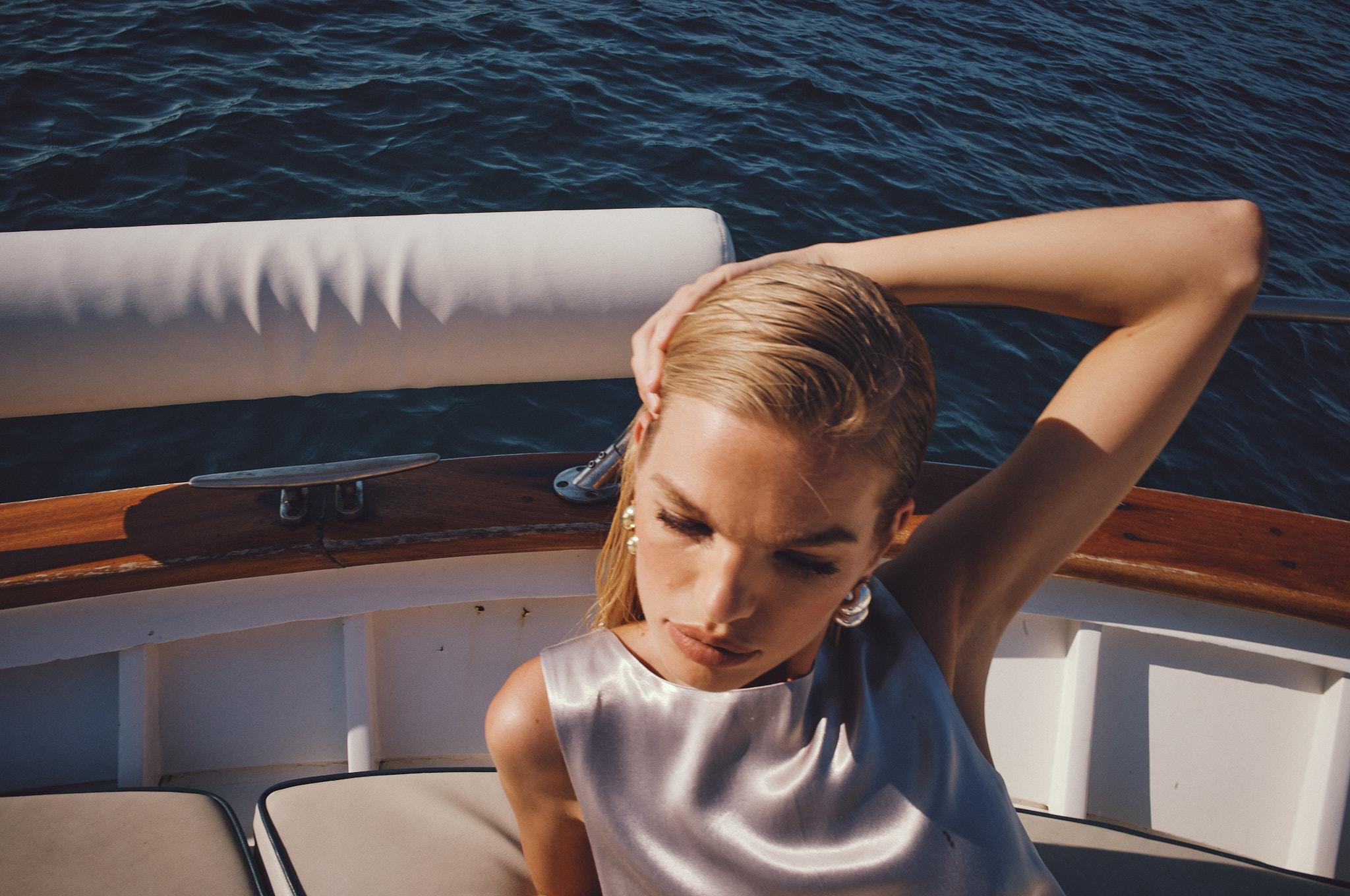 Woman with wet blonde hair, wearing a satin top and earrings, posing on a yacht with blue water visible in the background.