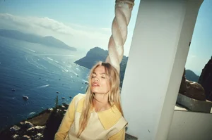Model Daphne Groeneveld leaning against a spiral column overlooking a scenic blue ocean coastline in a yellow sweater and cream vest.