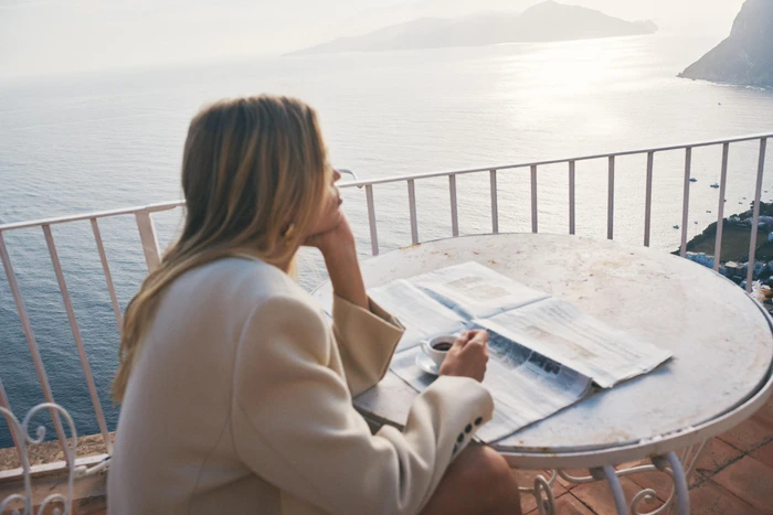 Daphne Groeneveld wearing a beige blazer seated at an outdoor cafe table with coffee and a newspaper overlooking the ocean.