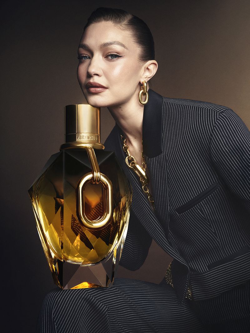Blonde woman in pinstripe suit poses with gold perfume bottle against brown gradient backdrop, gold jewelry, beauty, and fashion.