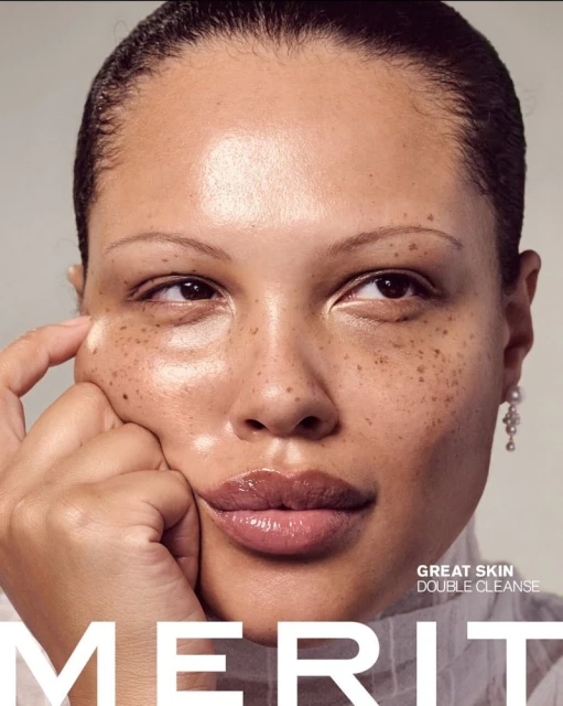 Close-up beauty shot of an adult female model with freckles, glossy lips, and pearl earrings promoting skincare, with Merit brand text.