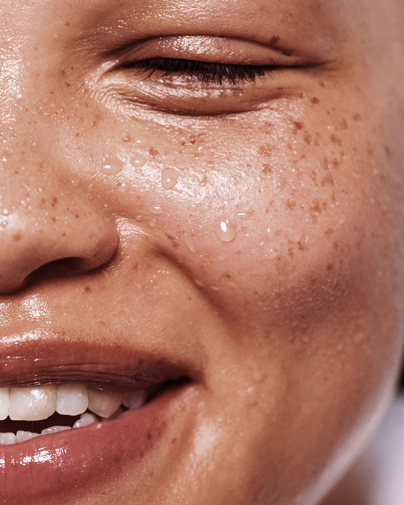Close-up beauty portrait of a smiling adult female with freckles and water droplets on her face. Glossy lips and warm lighting.