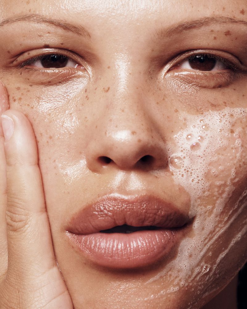 Close-up of a woman with freckles washing her face with soap, highlighting skincare and beauty in a studio setting.