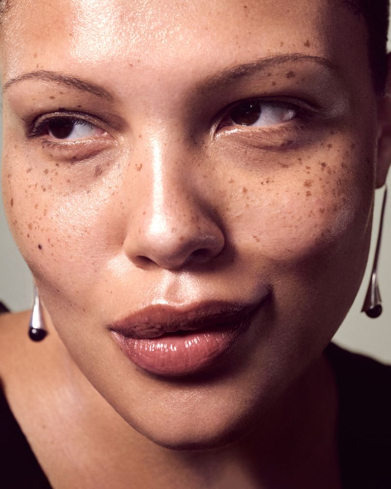 Close-up portrait of an adult female with freckles, teardrop earrings, and dark top, looking sideways in a brightly lit indoor setting.