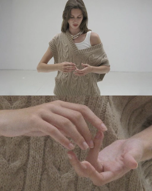 Woman in a sleeveless cable-knit sweater, hands in frame, posing in a neutral setting with soft lighting, showcasing fashion and style.