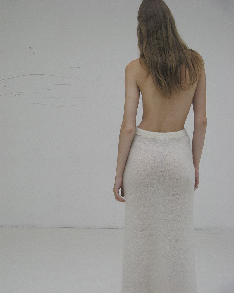 Back view of an adult woman with long brown hair wearing a long, textured white skirt in a minimalist indoor setting.