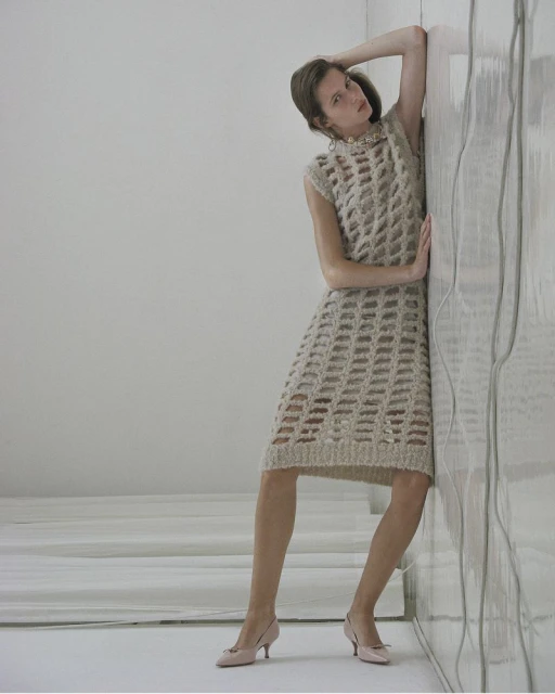 A woman in a beige open-weave dress and pumps leans against a textured wall in a minimalist, neutral-toned indoor studio fashion shot.
