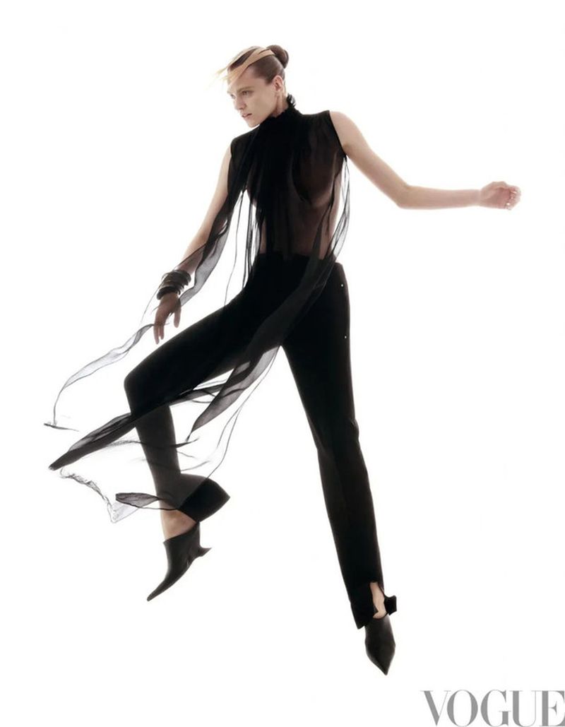 Fashion shot of androgynous model in motion wearing black sheer top and fitted pants against a white backdrop in a photo studio setting.