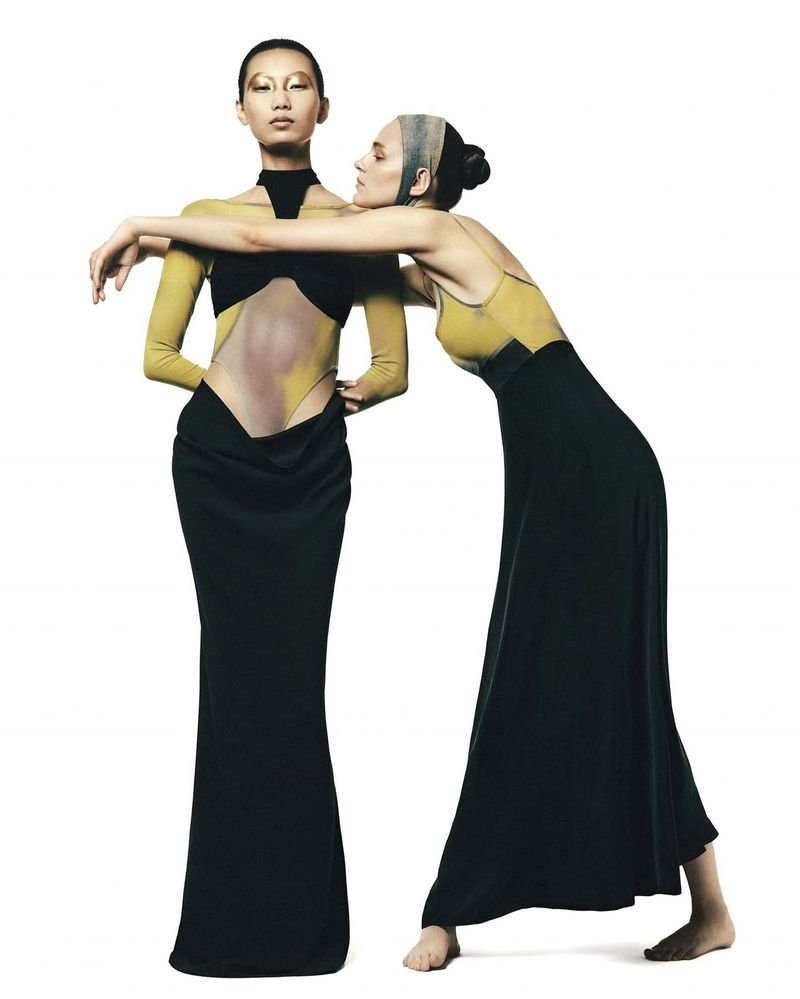 Two fashion models pose in a studio with gold eye makeup and yellow and black gowns, against a stark white background in a fashion editorial shot.