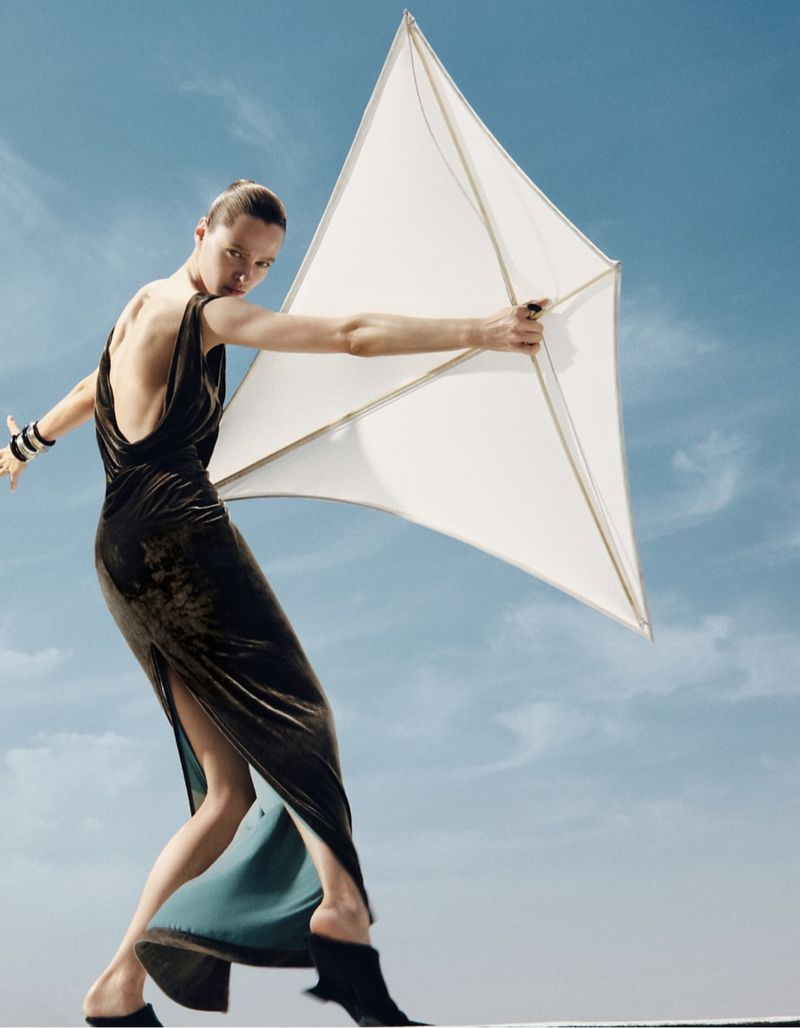 Fashion shot of a woman in a dark velvet dress holding a white kite against a blue sky. She is wearing black high heeled shoes.