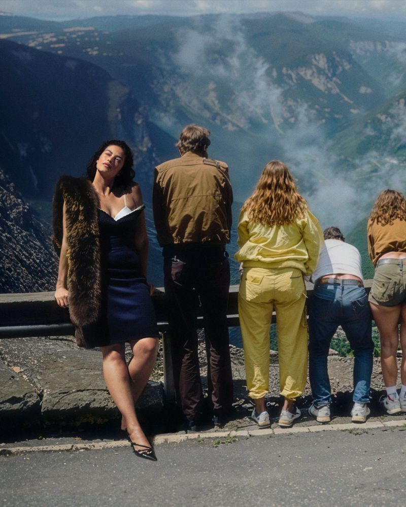 An adult woman poses in a stylish fur stole on a scenic overlook while a group of tourists look into the distance, enjoying a daytime view.