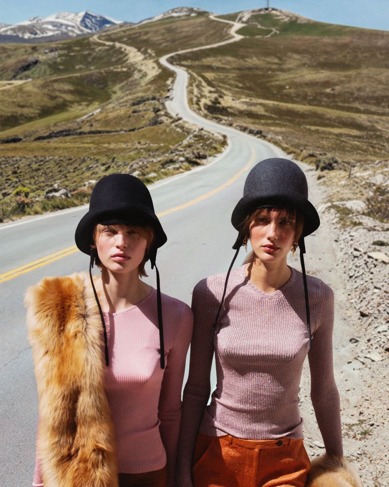 Two women pose on a remote road wearing hats and pink tops with mountains and a winding road in the background on a sunny day.