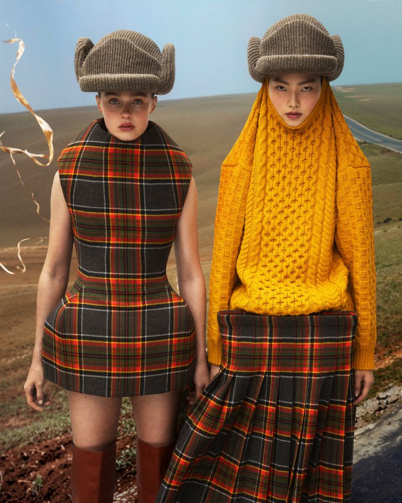 Two women in matching plaid outfits and hats pose in a tan field with a road in the background in a fashion editorial shot.