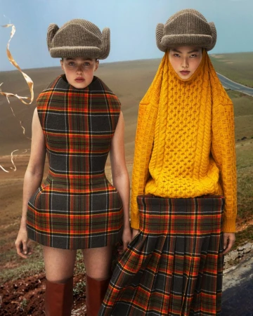 Two women in matching plaid outfits and hats pose in a tan field with a road in the background in a fashion editorial shot.