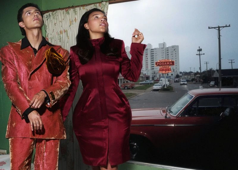 Two fashion models in red outfits pose indoors with a street view outside the window, showcasing an old car and building signs.
