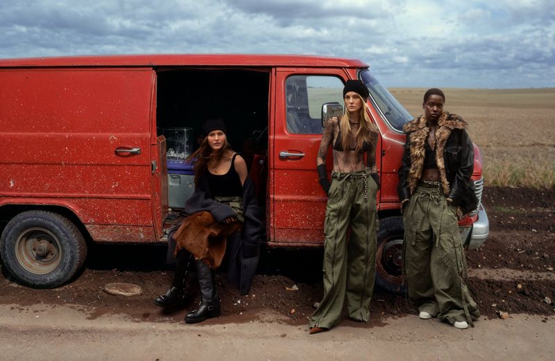 Three models pose in wide olive pants and crop tops in front of an open red van on a dirt road under a cloudy sky. 