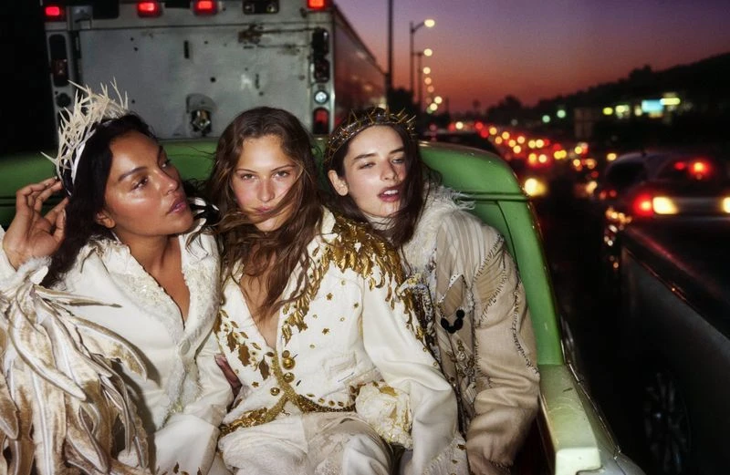 Three women in embellished white jackets and headpieces sit in a green car against a backdrop of city lights at dusk, creating an editorial fashion image.