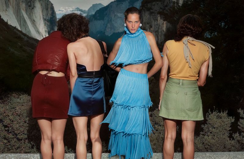 Four women wearing fashionable mini skirts and tops pose against a backdrop featuring mountains, creating a fashion-forward outdoor setting.