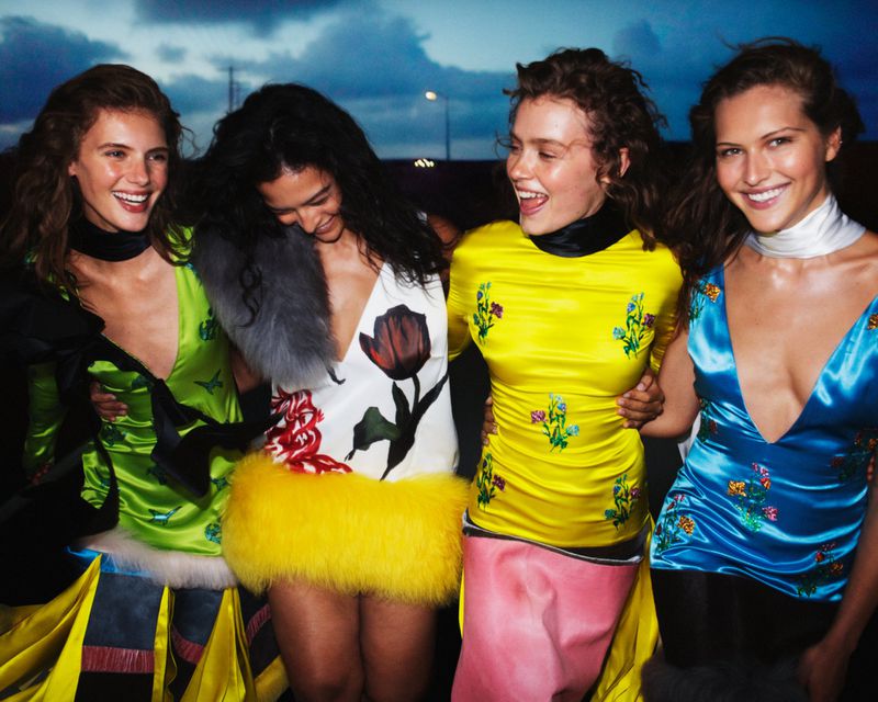 Four adult women pose outdoors, wearing colourful mini dresses, smiling and embracing in an eye-level medium shot under evening lighting.