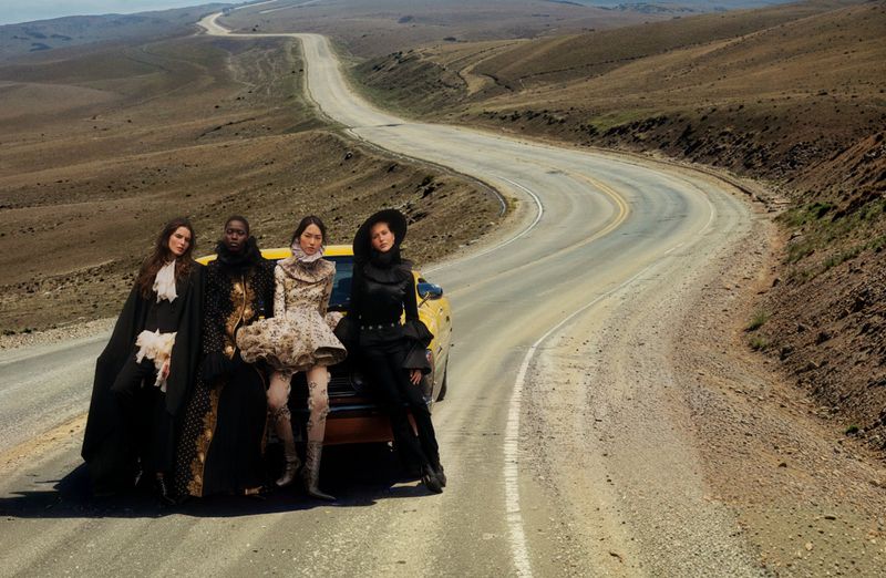 Four fashion models pose in couture dresses on a rural highway with a yellow car, set against a backdrop of rolling hills and sky.