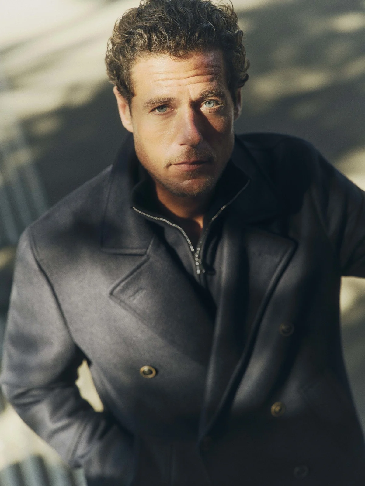 Portrait of a blue-eyed adult man with curly brown hair wearing a dark peacoat, shot from above, with natural outdoor lighting.