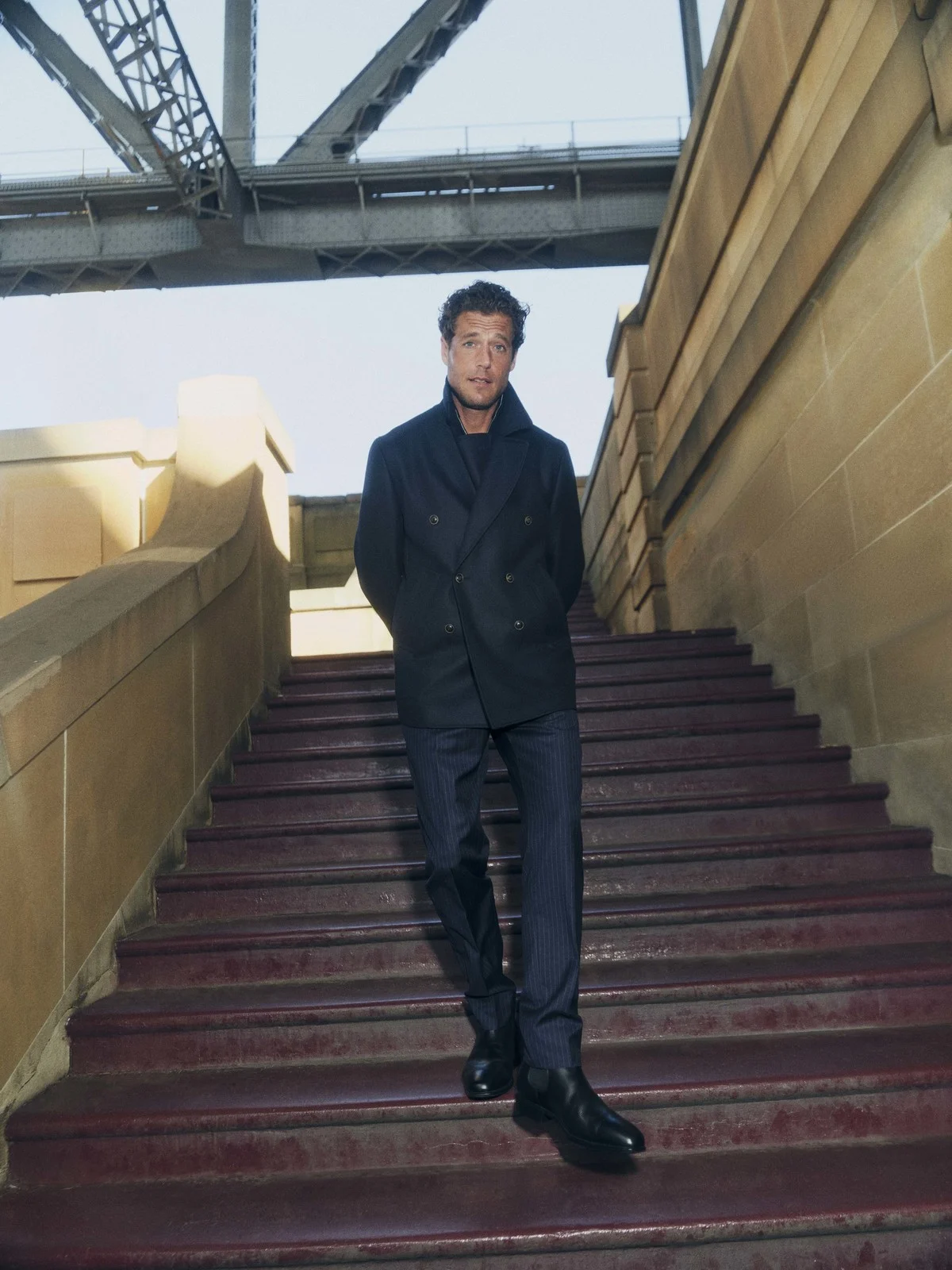Mature adult man in stylish black pea coat poses on stairs under bridge. Wearing pinstripe pants and boots, looking at the camera.
